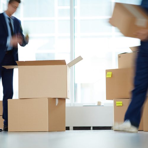 Businessman standing over open box by window of office center while relocation service worker carrying packages