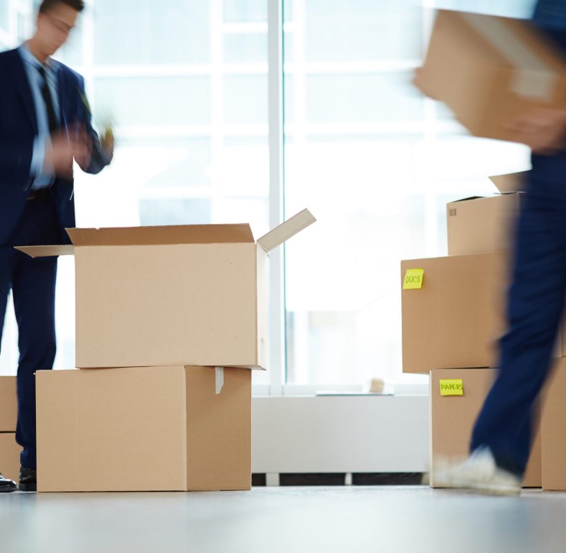 Businessman standing over open box by window of office center while relocation service worker carrying packages